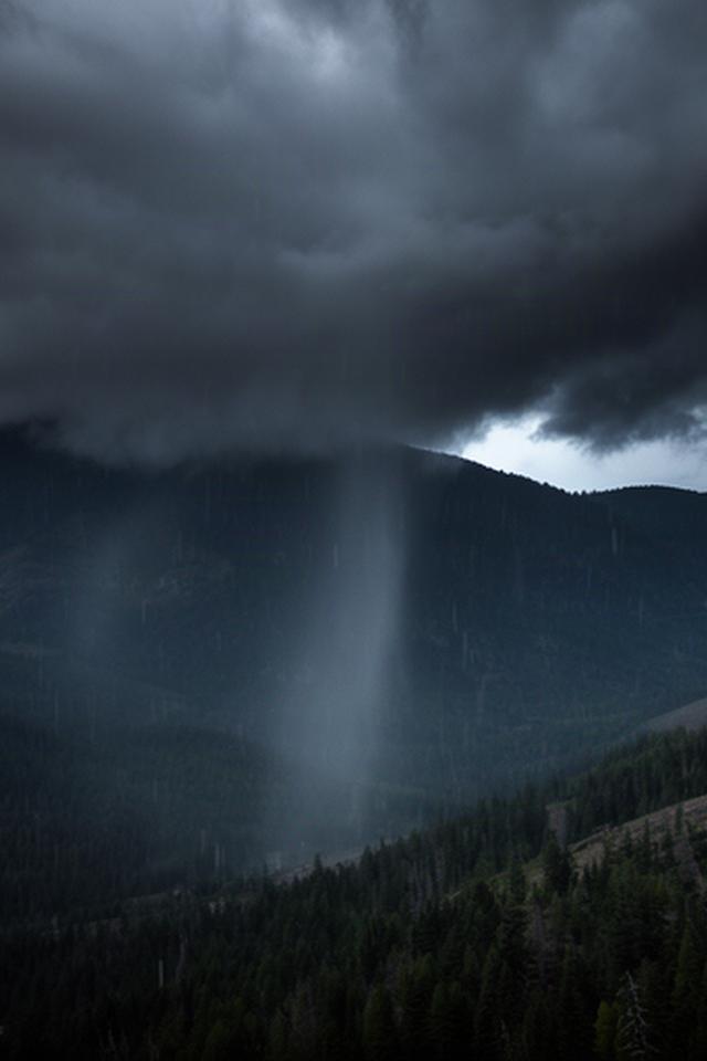Dunkle Gewitterwolken über einem Bergrücken, während Regen über einem bewaldeten Tal niedergeht